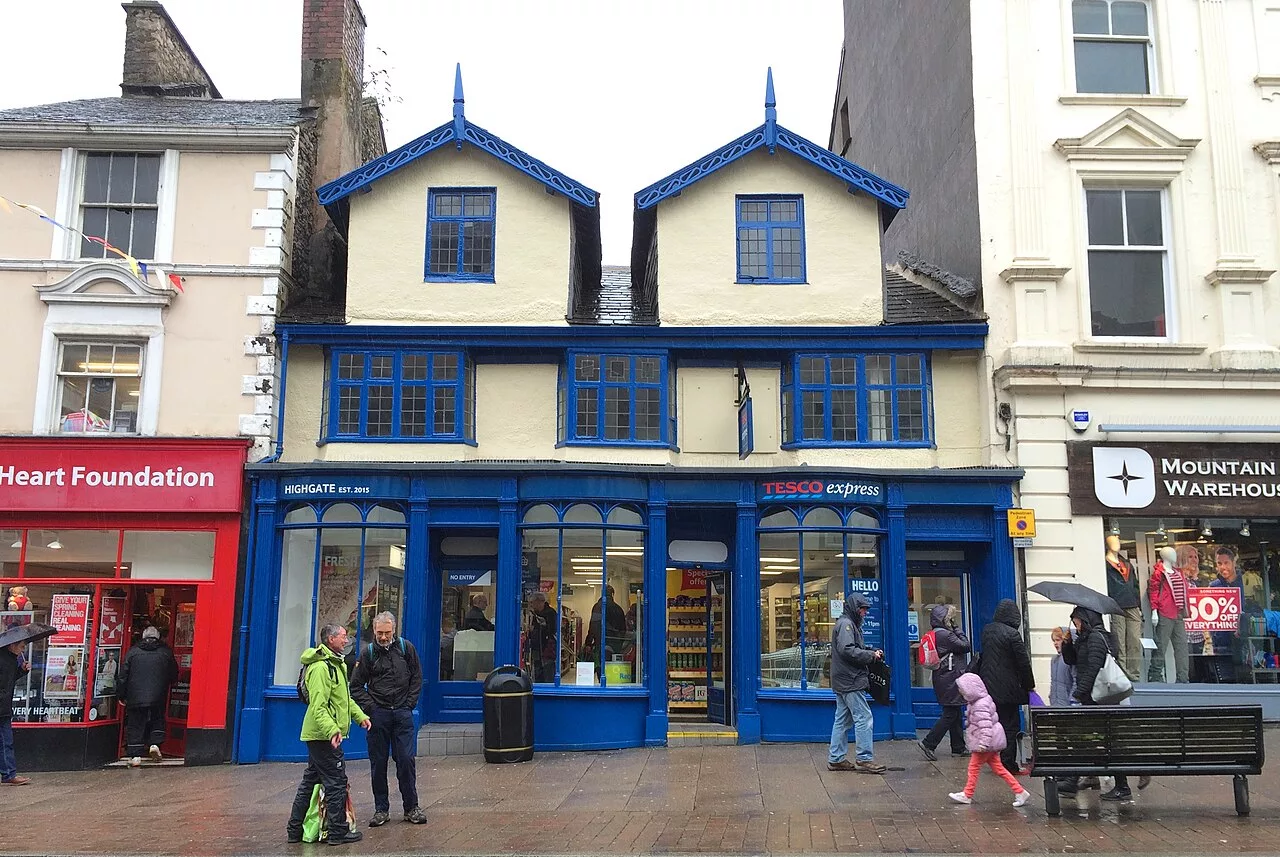 People walk past shops on a busy street. A blue and cream Tesco Express occupies a historic building with two pitched roofs. Nearby are the British Heart Foundation and Mountain Warehouse shops, as well as a local business offering House Removals In Kendal.